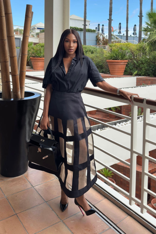 Woman in a black outfit with sheer skirt standing on a balcony with plants and a clear sky.