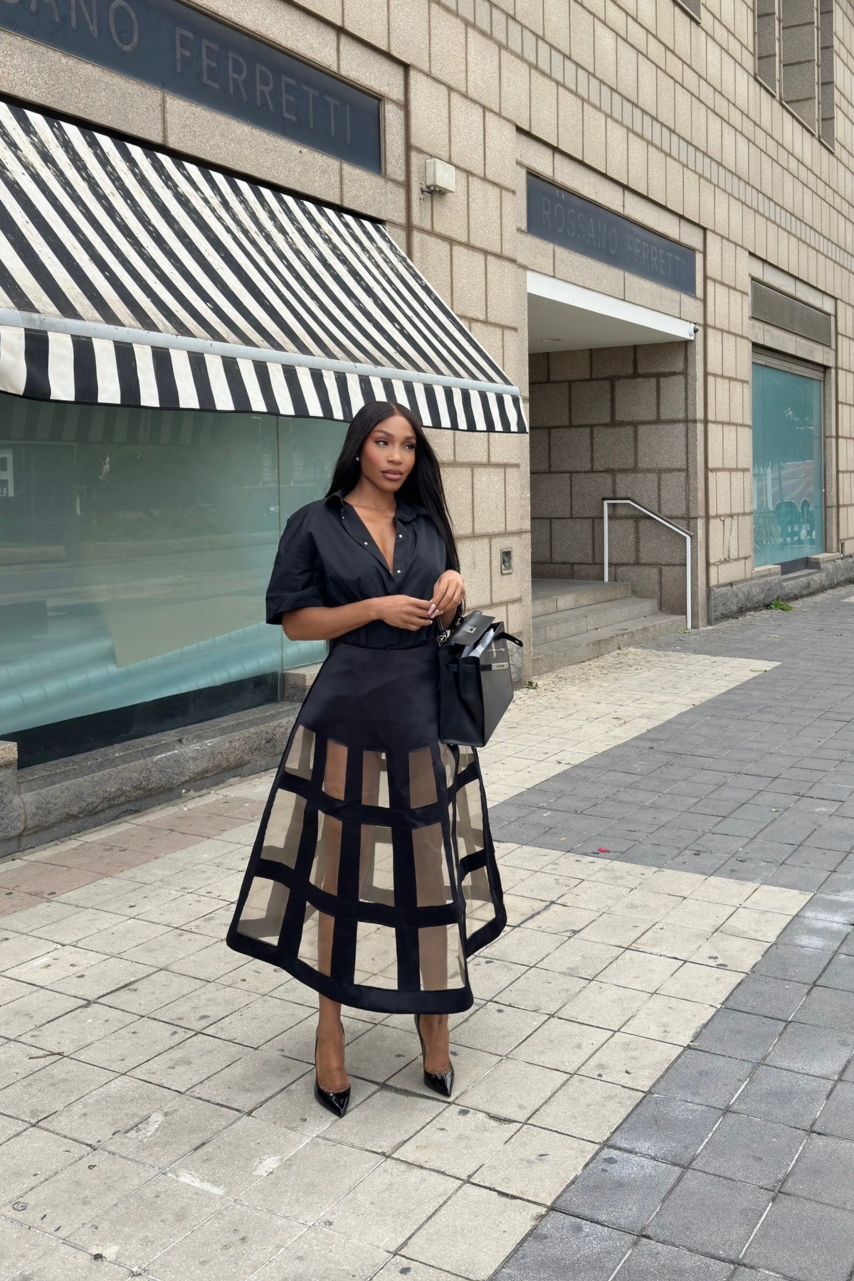 Woman in a black outfit standing on a city street with a building and striped awning in the background.
