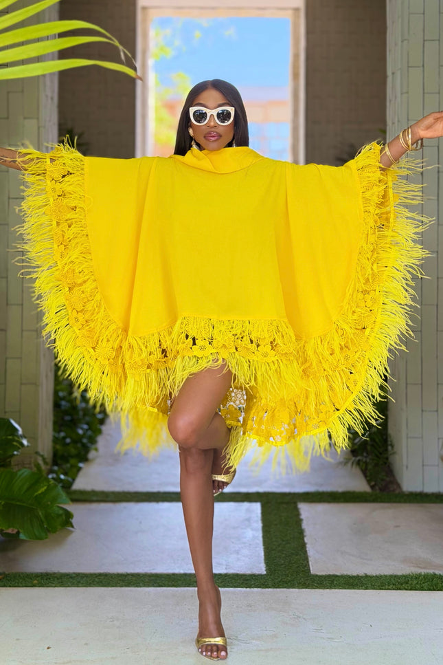 A woman modeling a bright yellow fringed poncho-style top with wide sleeves, photographed in an outdoor walkway with plants and a tiled wall background.