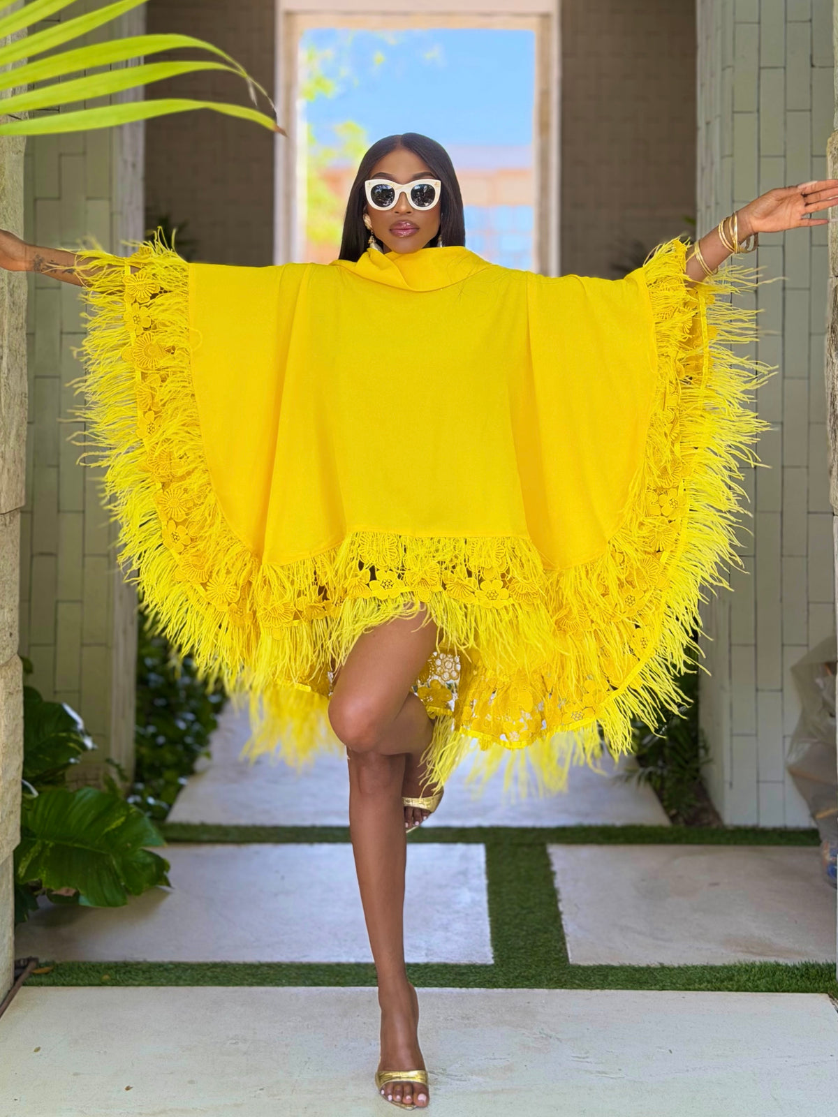 A woman modeling a bright yellow fringed poncho-style top with wide sleeves, photographed in an outdoor walkway with plants and a tiled wall background.